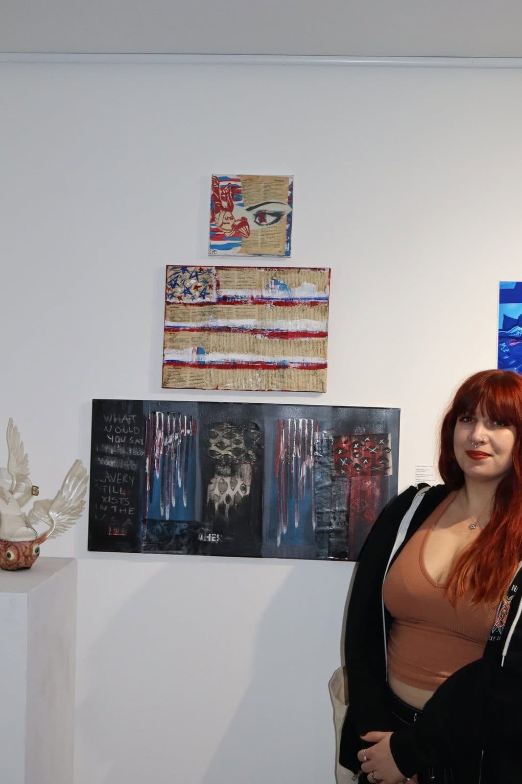 Jasmine Solei standing beside a wall of her mixed-media work at a gallery exhibition, including a stitched American flag piece and a row of dark panels