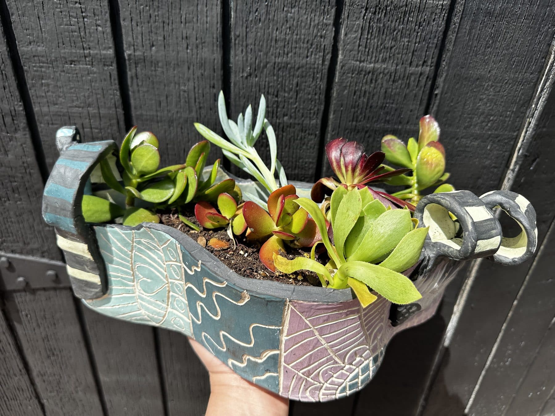 A hand-built ceramic planter shaped like an open, curling box with carved leaf patterns in slate blue and cream, overflowing with succulents, held up against a dark wood fence