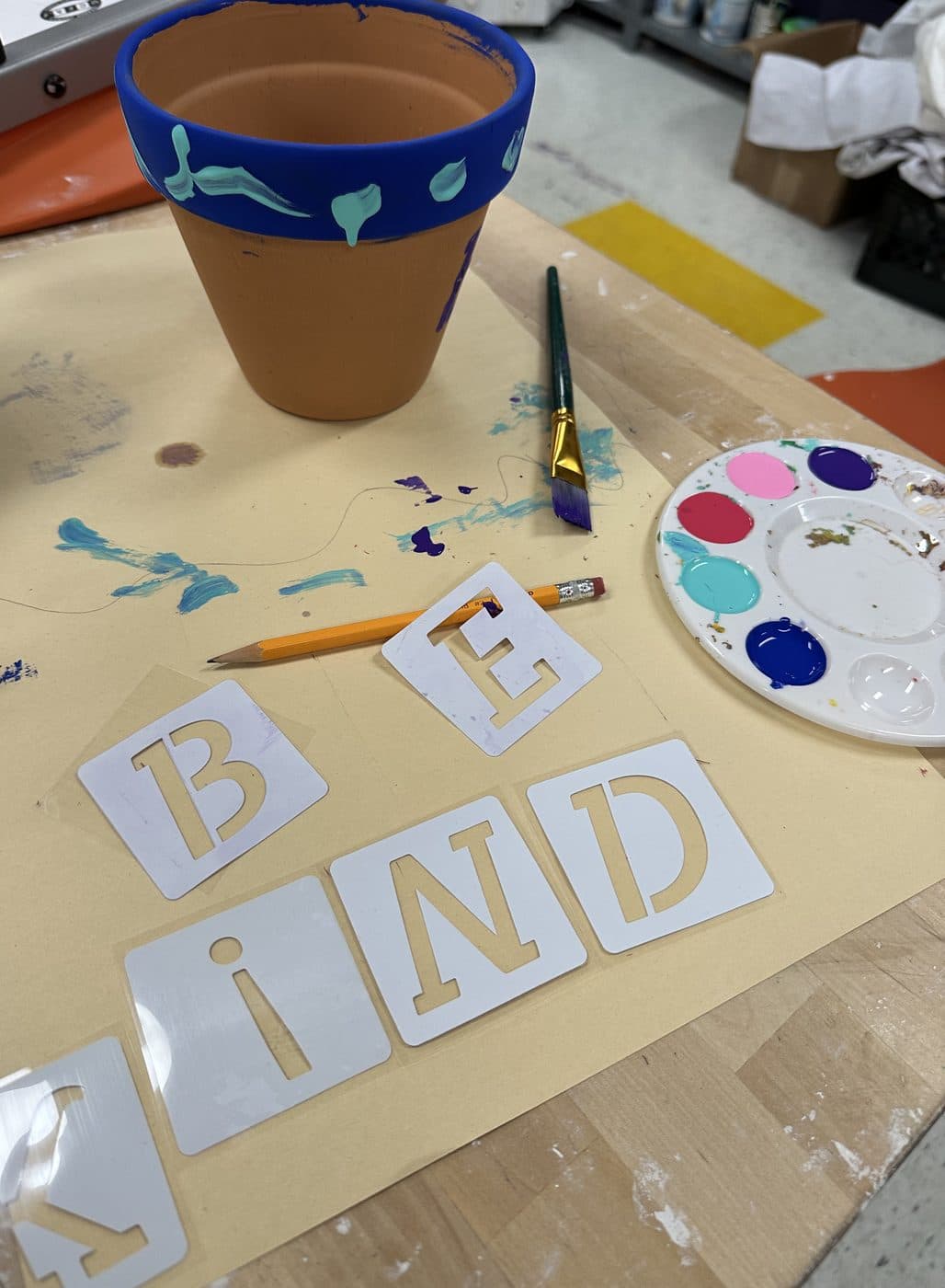 A work table with a freshly painted terracotta pot trimmed in blue, a paint palette, a brush, and letter stencils spelling “BE KIND”