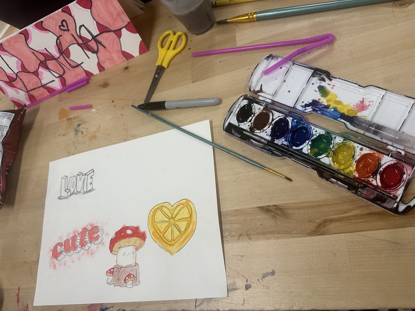 A wooden work table with an open watercolor set, brushes, scissors, and a page of small painted drawings, including a mushroom and a lemon-slice heart