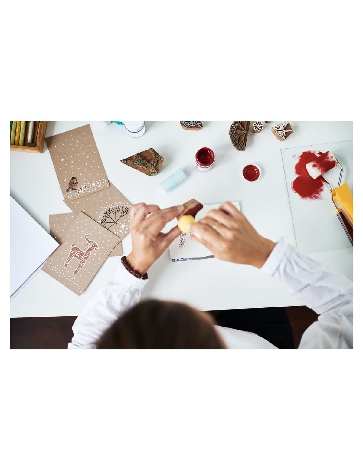 Overhead view of hands stamping a greeting card with a carved wood block, beside finished bird, deer, and snowflake cards and a red ink roller