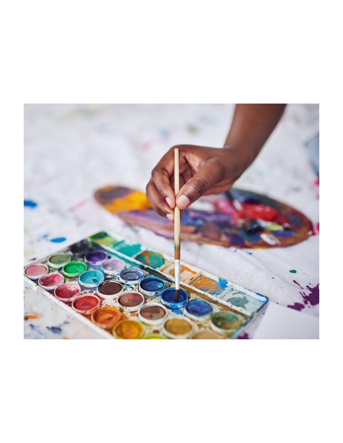 A hand dipping a paintbrush into a blue watercolor pan, with a paint-splattered cloth and a blurred palette in the background